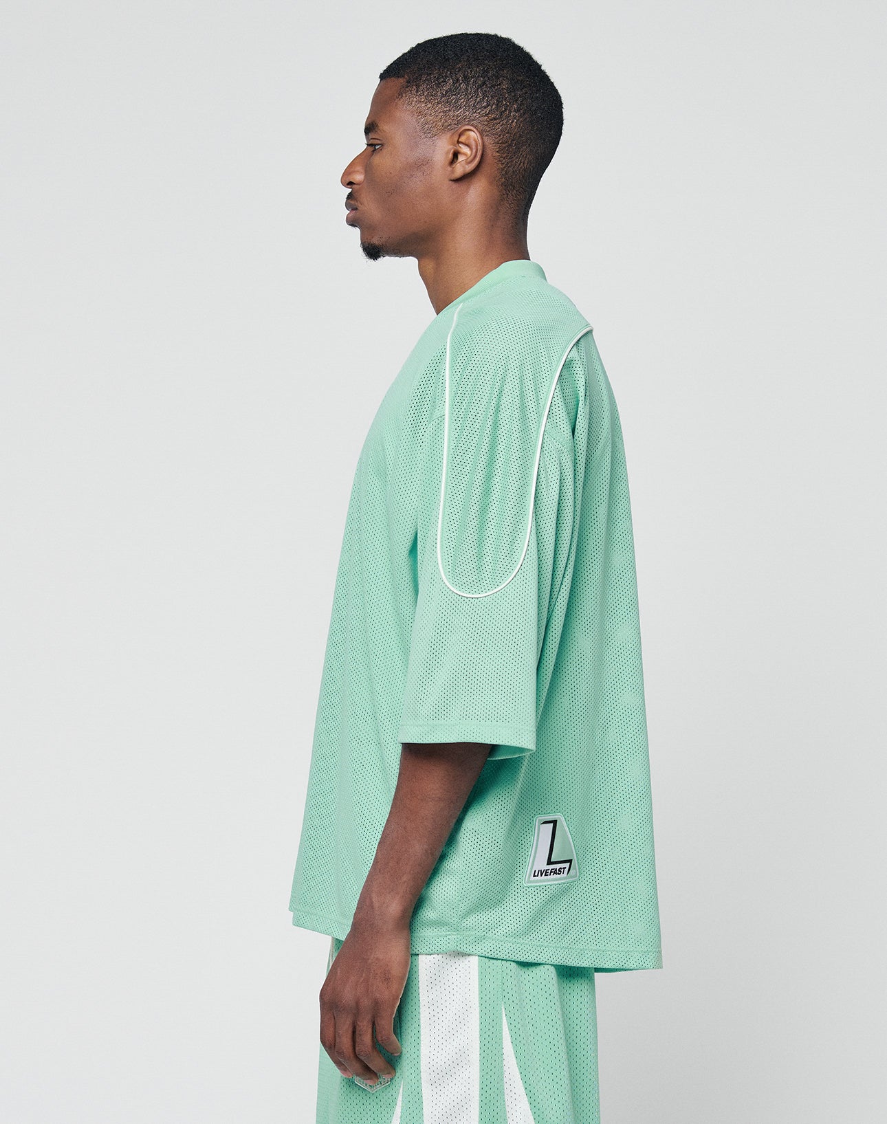 A man stands in profile wearing the LIVE FAST DIE YOUNG Basic Mesh Jersey and matching shorts in mint green with white contrast piping. The 100% polyester shirt features a small white and black patch on the lower back, set against a plain white background.