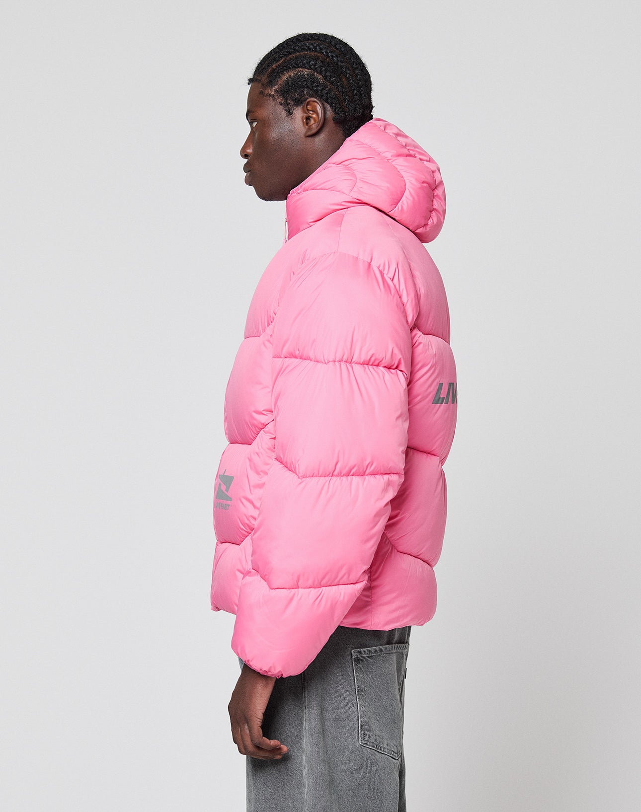 A person in profile wears the LIVE FAST DIE YOUNG Live Fast Puffer Jacket, a bright pink nylon jacket with an oversize fit and gray jeans. It features reflective gray text prints on the sleeve and back, set against a plain light background.