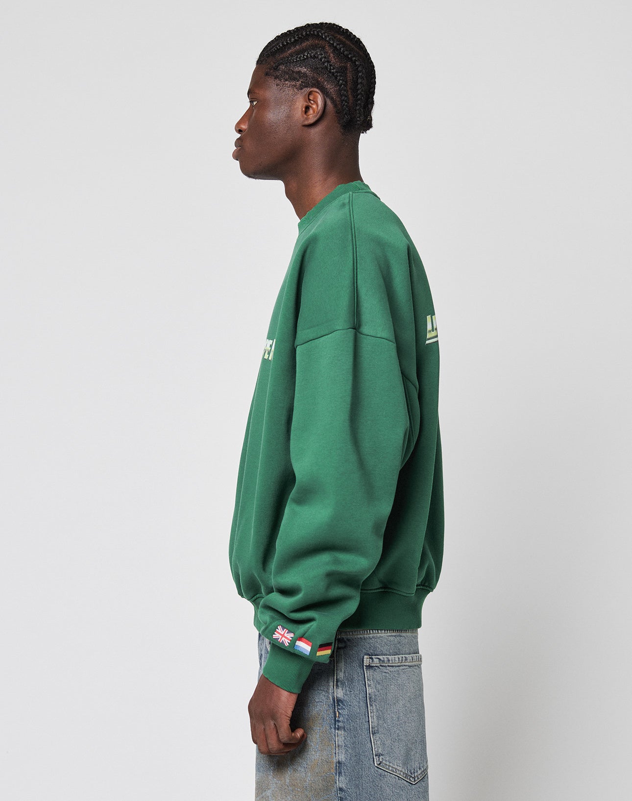 A person with braided hair is shown in profile wearing the LIVE FAST DIE YOUNG LFDY Europe Run Sweater in oversize fit green Baumwolle and faded blue jeans, with small flag patches near the left wrist, against a plain light grey background.
