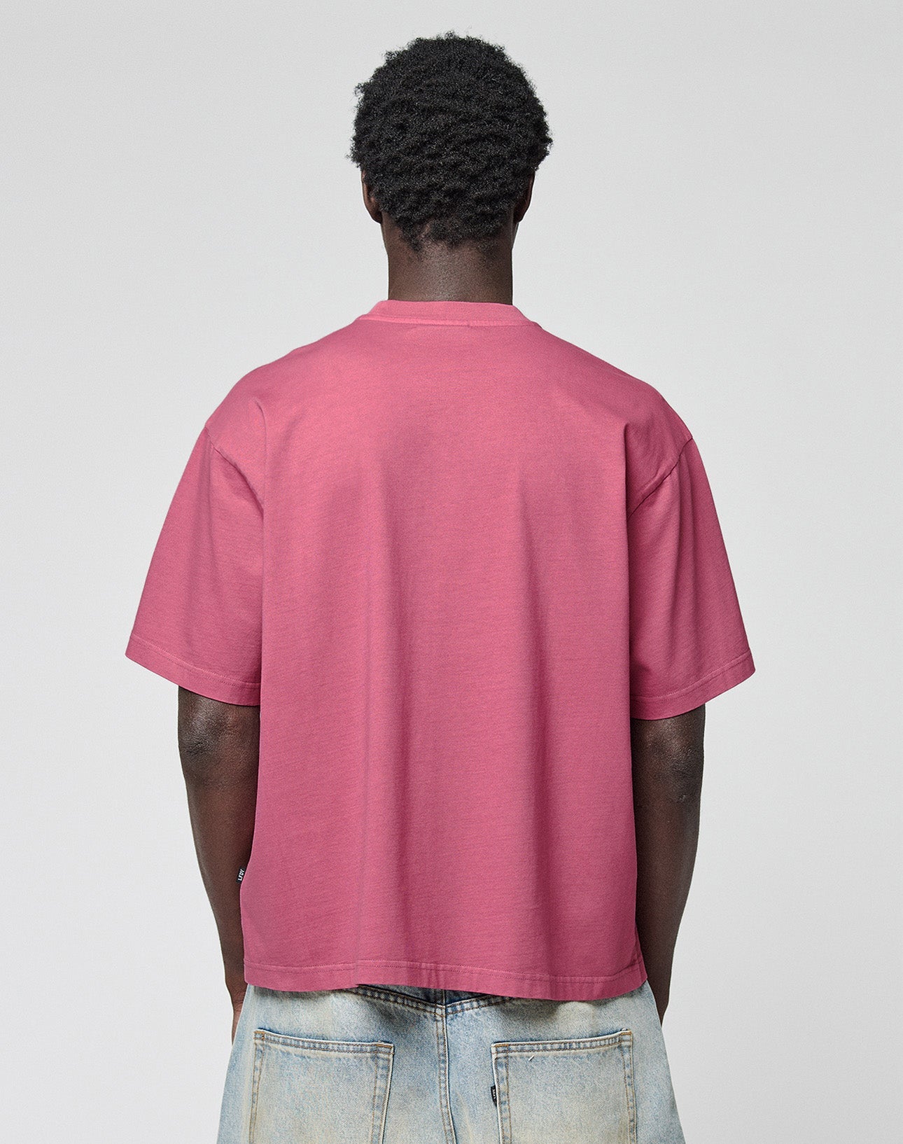 A person with short curly hair is seen from the back, wearing the oversized pink Live Fast LF Tee by LIVE FAST DIE YOUNG and light blue jeans against a plain light gray background.
