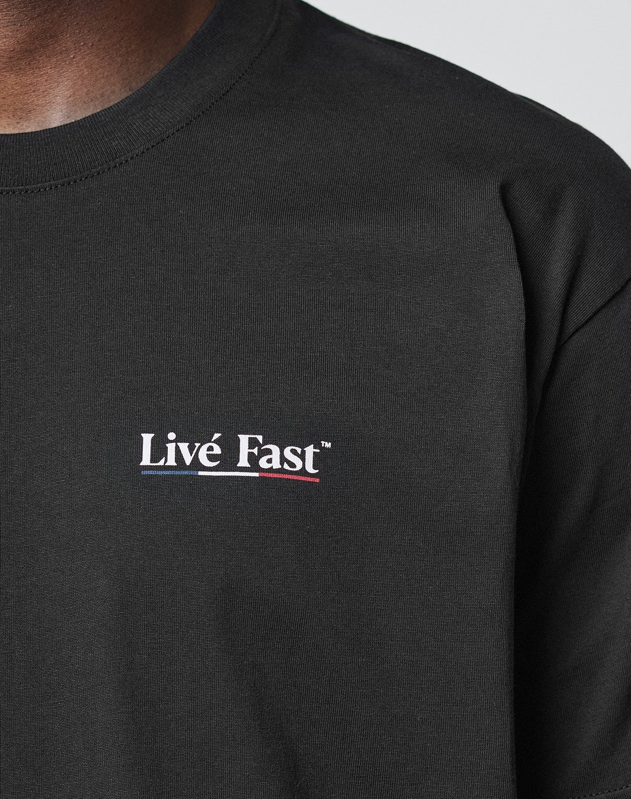 A close-up of a person wearing the Live Fast Paris Tee by LIVE FAST DIE YOUNG. This black, regular fit T-shirt features bold white Live Fast™ text on the chest, stylish front and back prints, and is made from 100% Baumwolle.