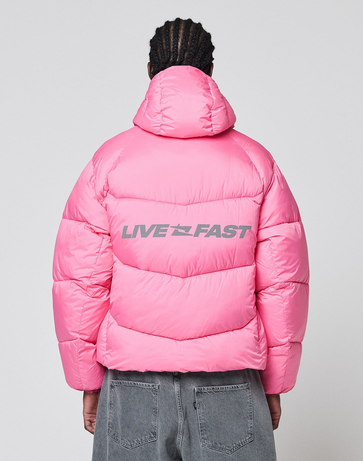 A person faces away, wearing the LIVE FAST DIE YOUNG Live Fast Puffer Jacket in bright pink Nylon with an oversized fit and hood. "LIVE FAST" is printed in bold gray reflective letters on the back. They pair it with faded gray jeans.