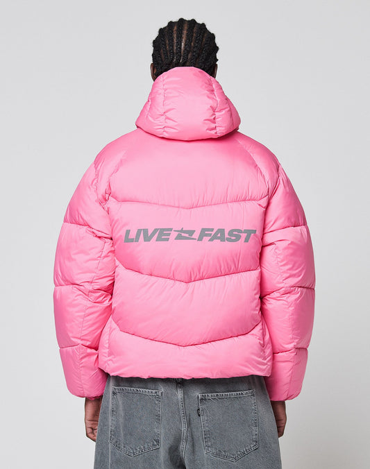 A person faces away, wearing the LIVE FAST DIE YOUNG Live Fast Puffer Jacket in bright pink Nylon with an oversized fit and hood. "LIVE FAST" is printed in bold gray reflective letters on the back. They pair it with faded gray jeans.