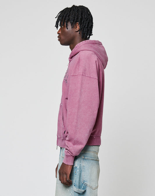 A person with short, textured black hair stands sideways in a LIVE FAST DIE YOUNG Live Fast Rivet Hooded Zip (faded mauve) and light blue paint-splattered jeans, against a plain light gray background.