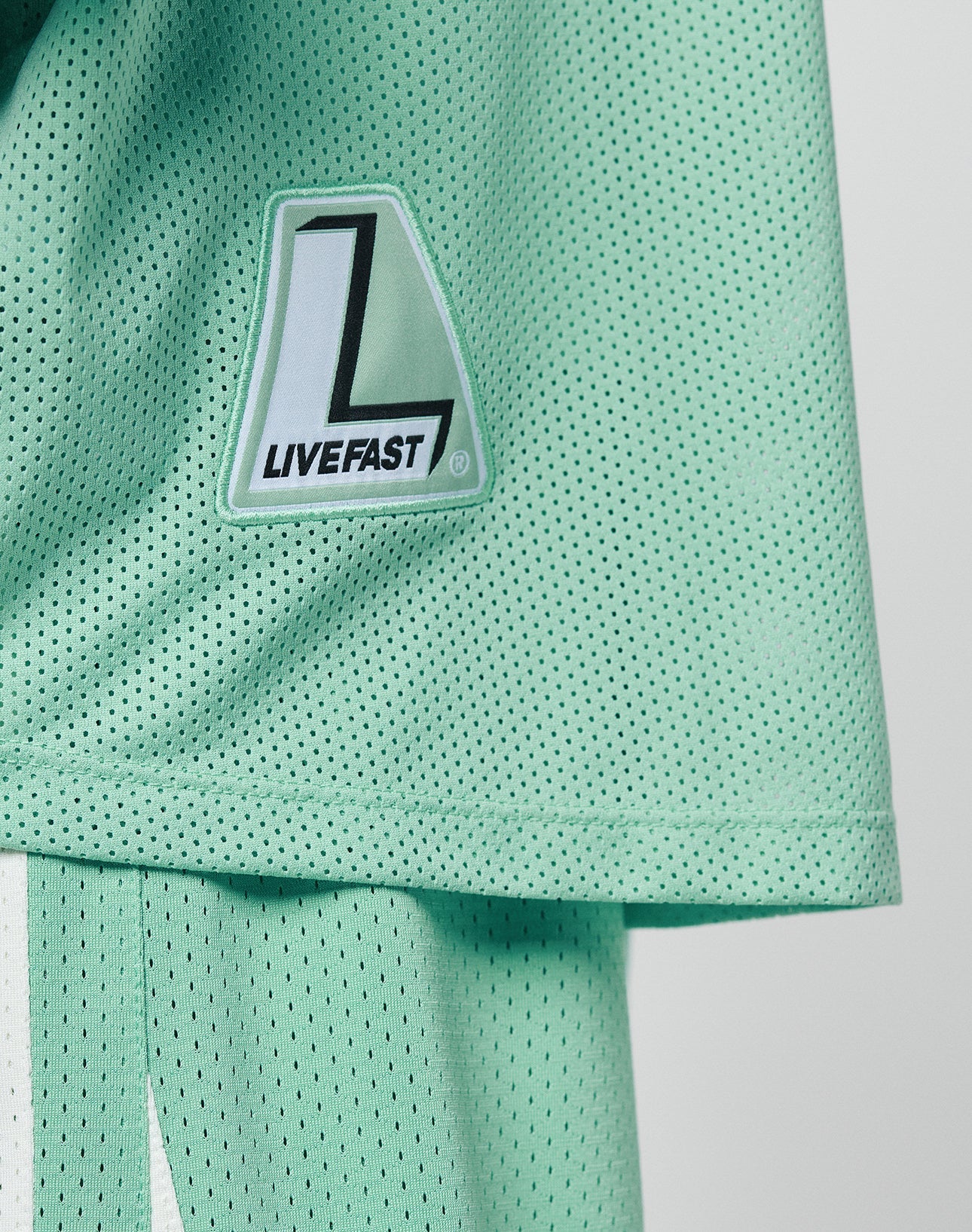 A close-up of the mint green Basic Mesh Jersey by LIVE FAST DIE YOUNG, featuring 100% polyester mesh and a patch with a large L and LIVEFAST. Matching shorts with a white stripe and contrast piping are partially visible.