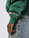 A person wears the LFDY Europe Run Sweater by LIVE FAST DIE YOUNG—an oversize-fit green sweatshirt with embroidered UK, Netherlands, and Germany flags on the sleeve—crafted from a soft Baumwolle-Polyester blend, paired with blue jeans.