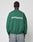 A person with braided hair, facing away, wears the LIVE FAST DIE YOUNG LFDY Europe Run Sweater in green with bold white LIVE FAST RACE AM text on the back, paired with light blue jeans.