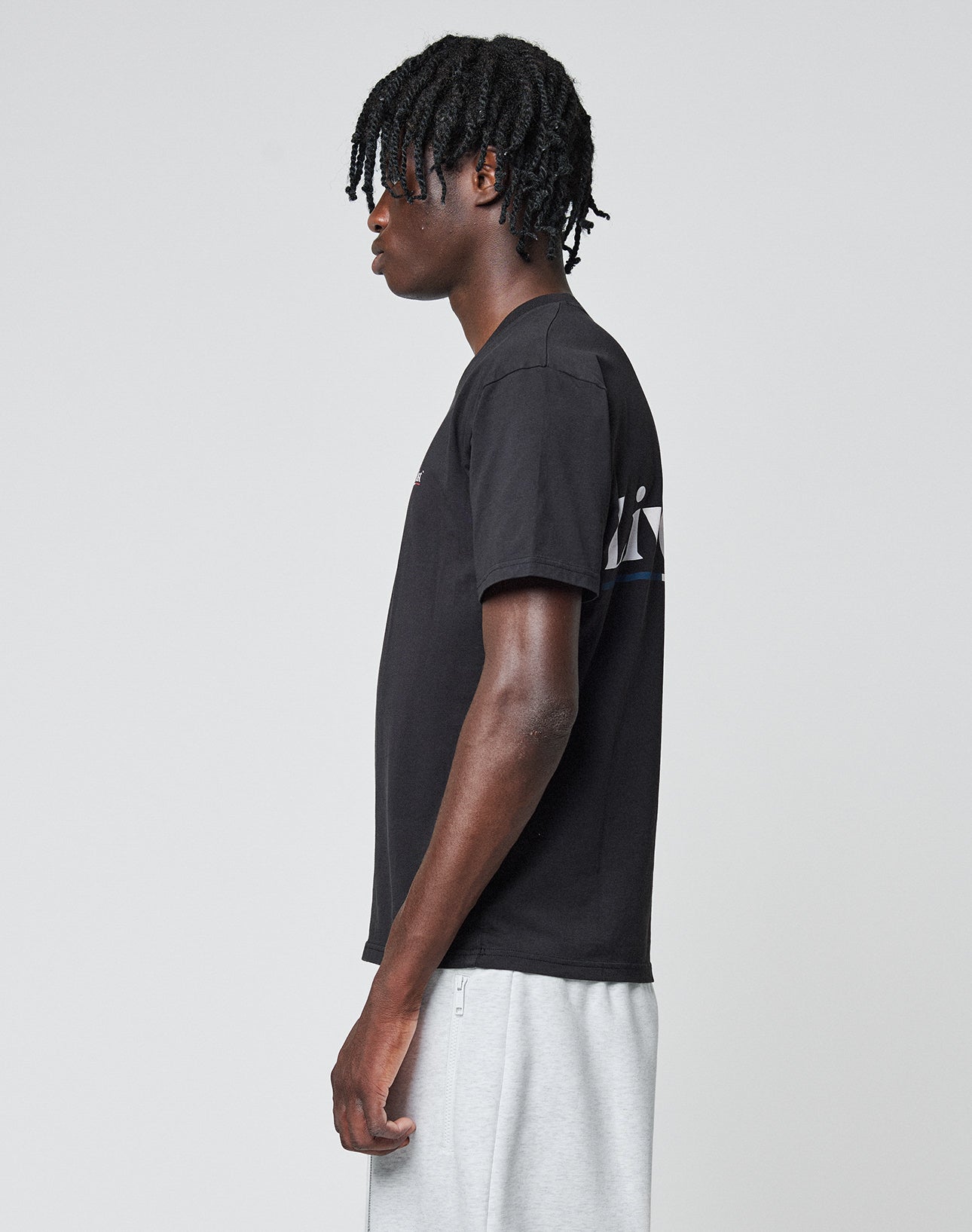 A person with medium-length, twisted hair stands in profile wearing the LIVE FAST DIE YOUNG Live Fast Paris Tee and light gray shorts against a plain, light background.