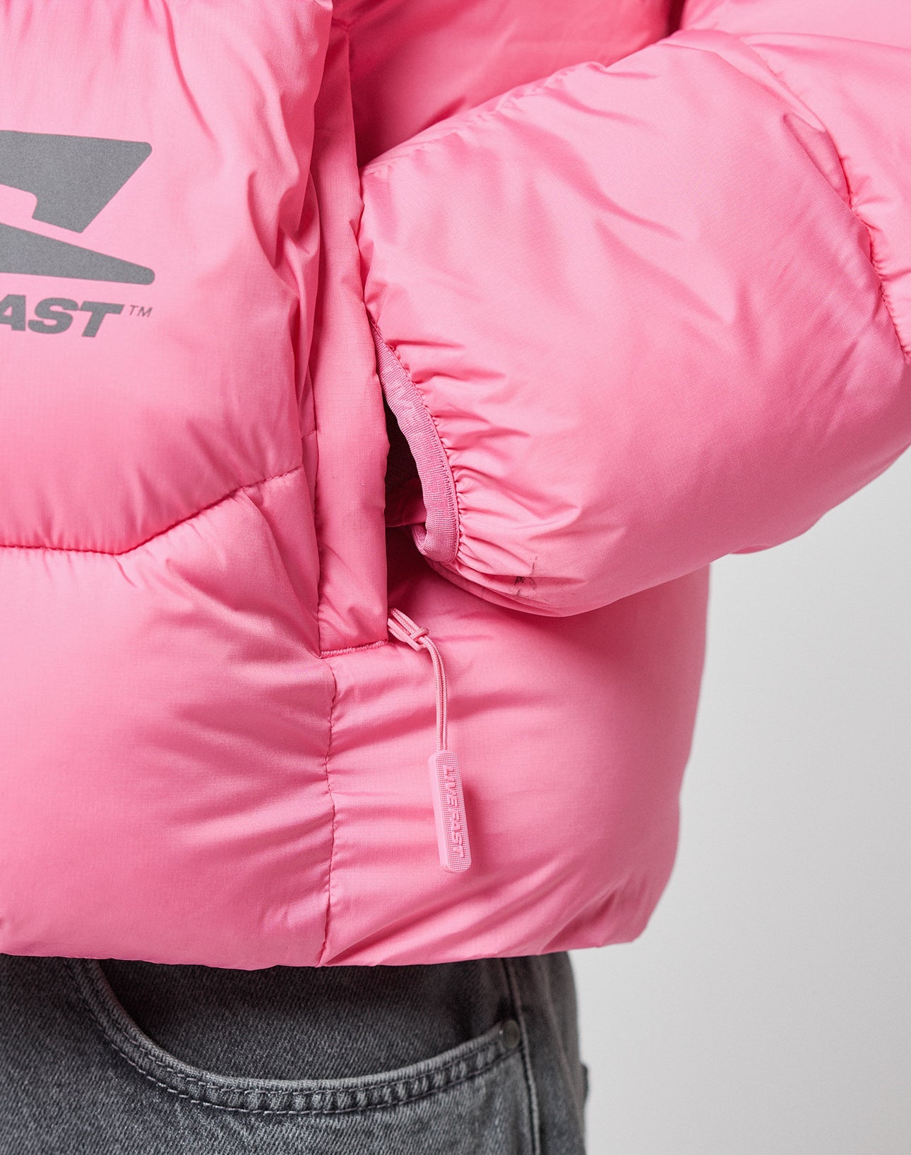 Close-up of a person wearing the LIVE FAST DIE YOUNG Live Fast Puffer Jacket in bright pink nylon with an oversize fit, visible zipper pull, and sleeve logo, paired with gray denim pants and a hand tucked into the jacket pocket.