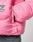 Close-up of a person wearing the LIVE FAST DIE YOUNG Live Fast Puffer Jacket in bright pink nylon with an oversize fit, visible zipper pull, and sleeve logo, paired with gray denim pants and a hand tucked into the jacket pocket.