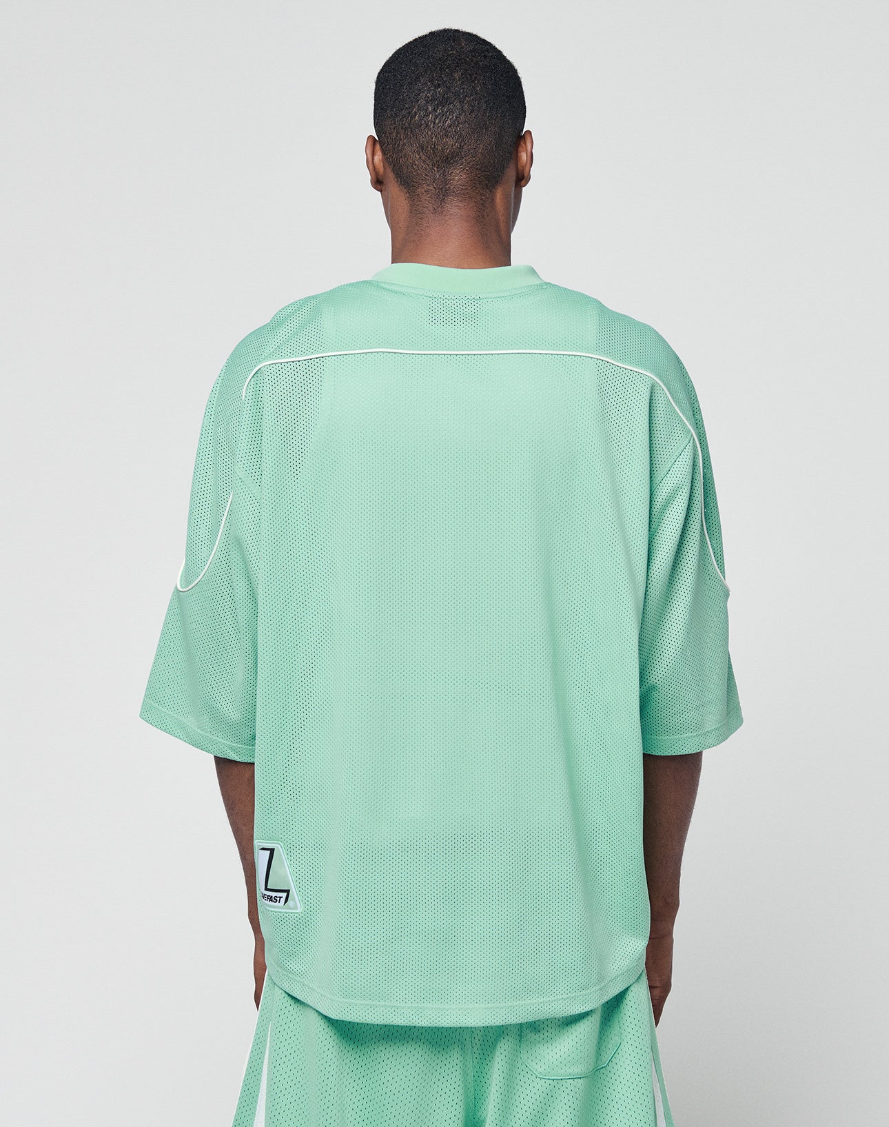 A person faces away, wearing the LIVE FAST DIE YOUNG Basic Mesh Jersey and matching shorts in light green 100% polyester with white contrast piping and a small Z patch on the lower back. The background is plain white.