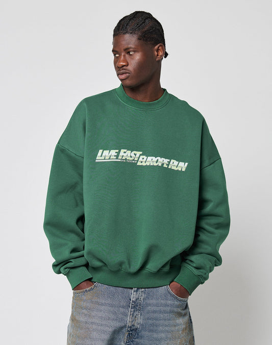 A person wears the LFDY Europe Run Sweater by LIVE FAST DIE YOUNG—an oversize fit green Baumwolle sweatshirt with LIVE FAST BEFORE RUN on the front—paired with blue jeans, standing against a plain light background, hands in pockets.
