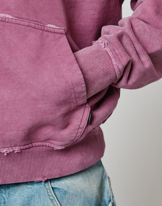 A person wearing a Live Fast Rivet Hooded Zip sweatshirt by LIVE FAST DIE YOUNG—faded mauve with distressed details and handgefertigter Sprayeffekt—paired with light blue denim jeans, right hand in the 100% Baumwolle sweatshirt pocket.