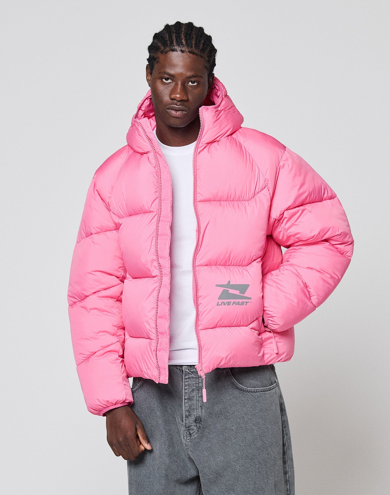 A man wears the bright pink LIVE FAST DIE YOUNG Live Fast Puffer Jacket with an oversize fit and reflective "LIVE FAST" print near the pocket, standing against a light gray background over a white T-shirt.