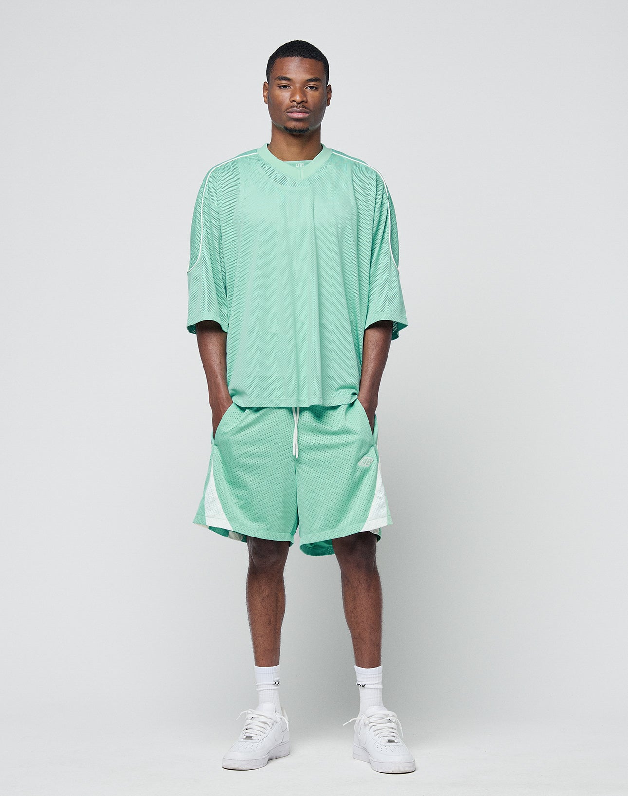 A man stands facing forward in a relaxed mint green Striped Mesh Shorts set by LIVE FAST DIE YOUNG, featuring white side panels and a discreet logo patch. He has his hands in his pockets, wears white sneakers, against a plain light gray background.