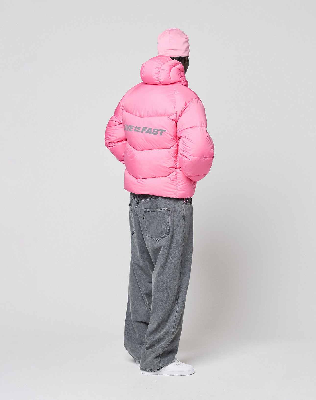 A person stands facing away in a bright pink Live Fast Puffer Jacket by LIVE FAST DIE YOUNG, featuring reflective "NEEFAST" print on the back, paired with a pink cap, loose gray jeans, and white sneakers against a plain white background.