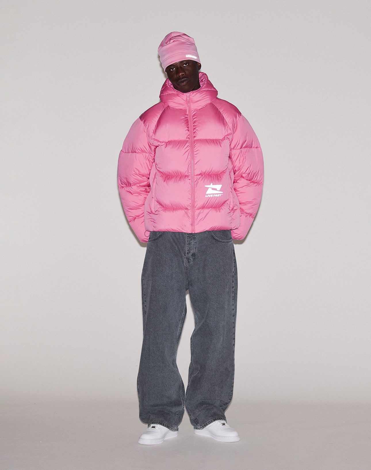 A person stands against a plain background wearing the bright pink Live Fast Puffer Jacket by LIVE FAST DIE YOUNG, a matching pink beanie, loose gray jeans, and white sneakers.