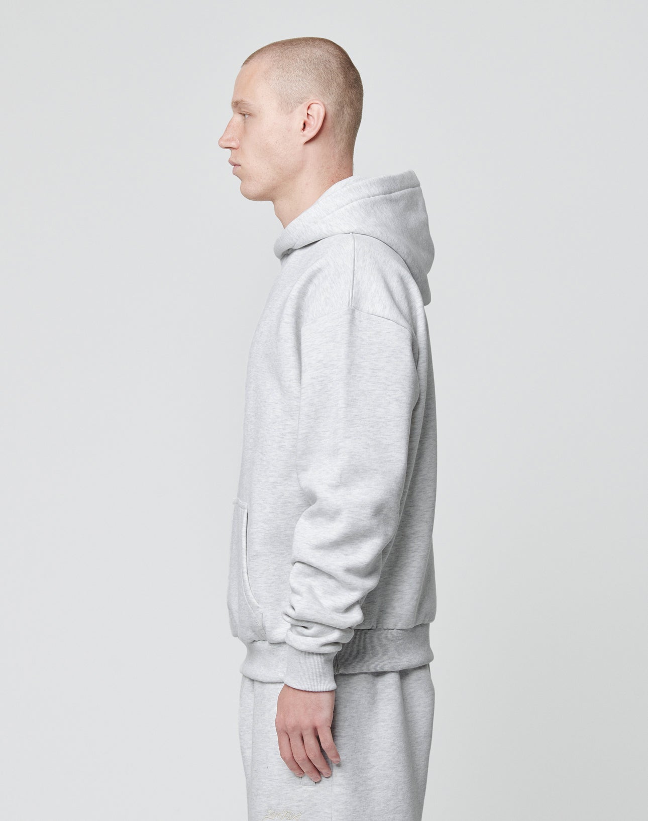 A person with a shaved head stands in profile, facing left, wearing the LIVE FAST DIE YOUNG Basic Blank Hooded sweatshirt and matching sweatpants in light gray Baumwolle-Polyester blend against a plain light background.