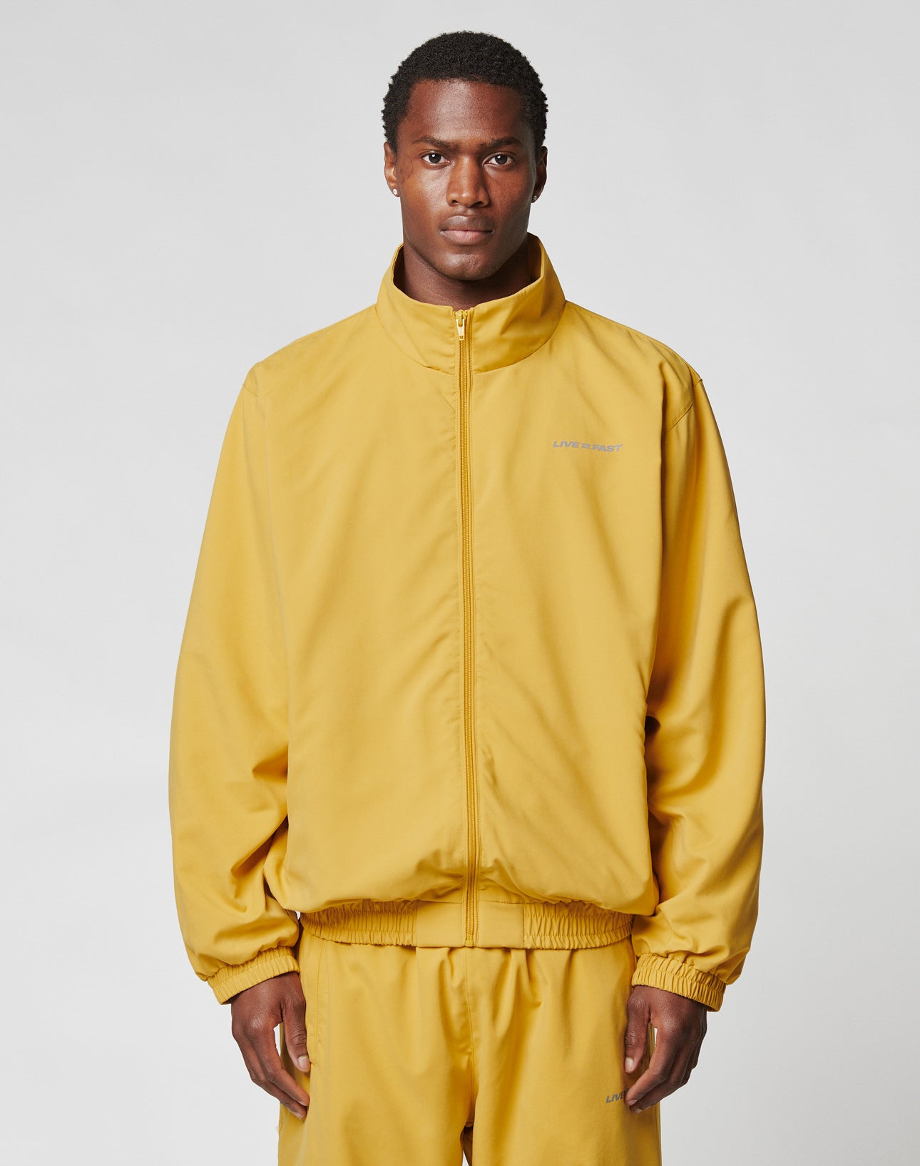 A man wears the LIVE FAST DIE YOUNG LFDY Trackjacket in an oversize fit, mustard yellow with reflective print, along with matching pants. He stands facing forward against a plain light gray background.