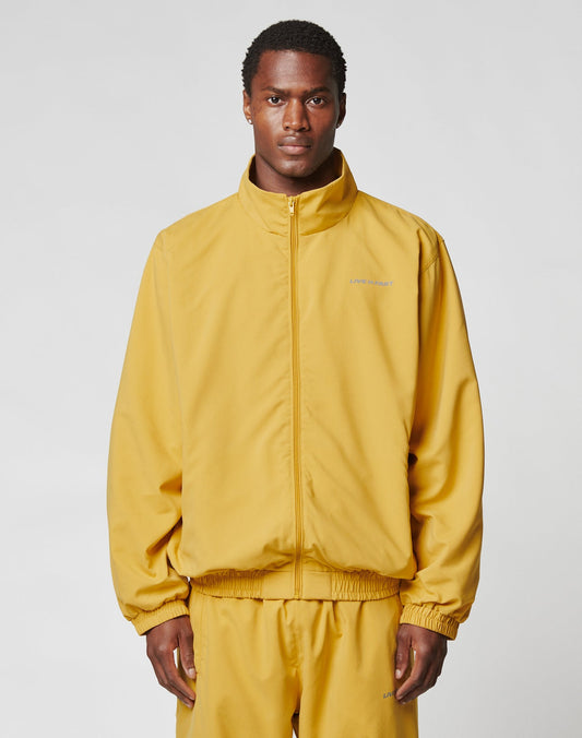 A man wears the LIVE FAST DIE YOUNG LFDY Trackjacket in an oversize fit, mustard yellow with reflective print, along with matching pants. He stands facing forward against a plain light gray background.