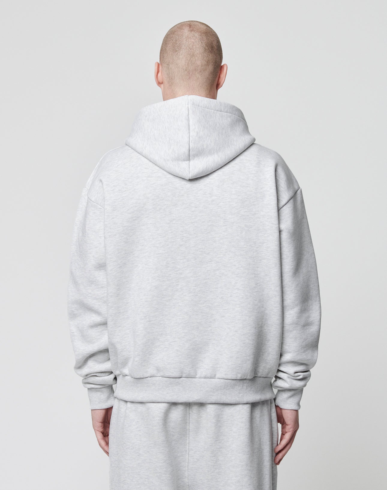 A person with a shaved head, seen from behind, wears an oversize fit light gray LIVE FAST DIE YOUNG Basic Blank Hooded sweatshirt and matching sweatpants made of soft Baumwolle against a plain light background.