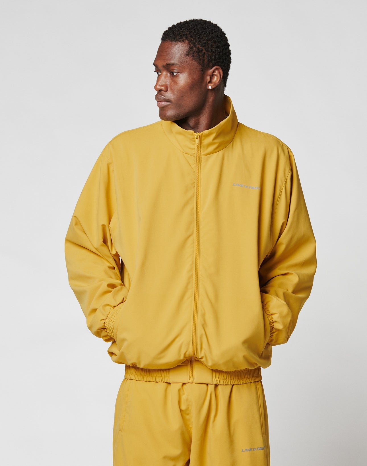 A man wearing the LIVE FAST DIE YOUNG LFDY Trackjacket in yellow with matching oversized pants stands with his hands in his pockets, looking to the side against a plain white background.