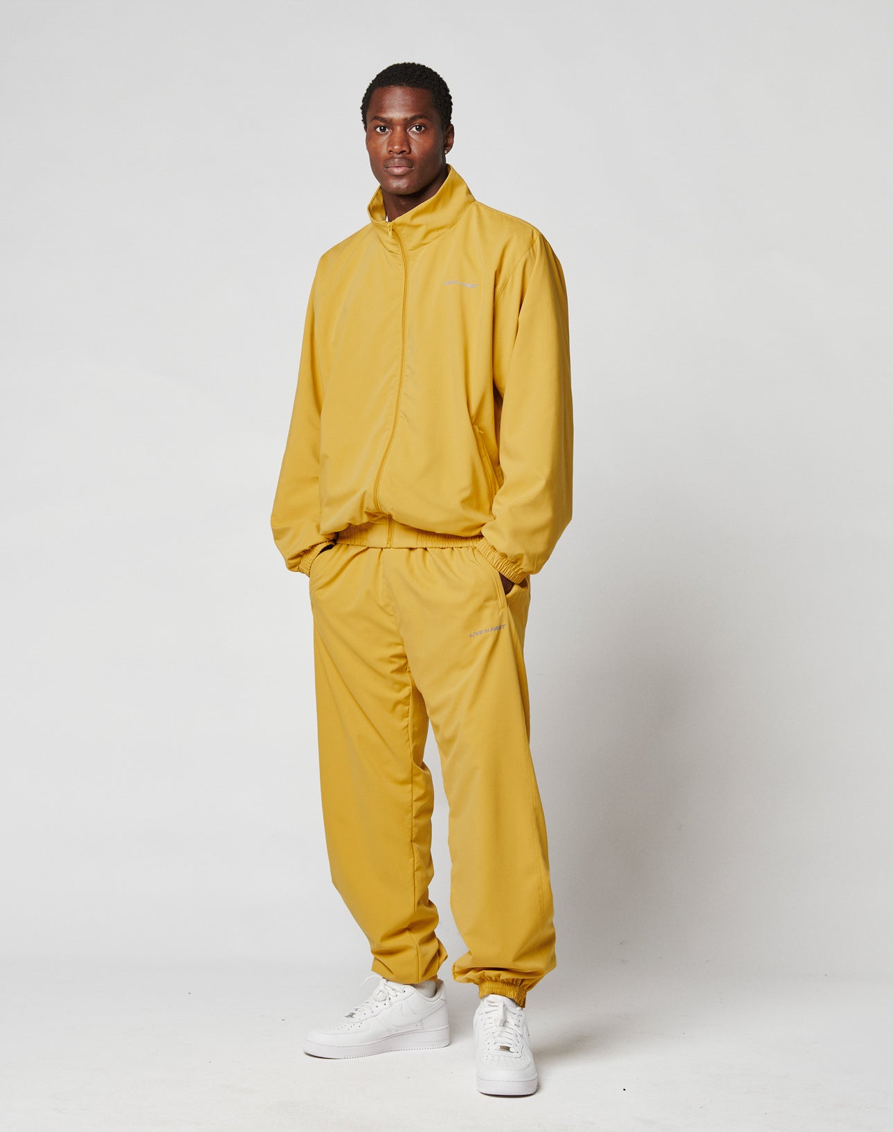 A man stands against a white background, hands in pockets, wearing LIVE FAST DIE YOUNG LFDY Trackpants—loose-fitting mustard yellow polyester with Elastikbund and reflective print—confidently looking at the camera.
