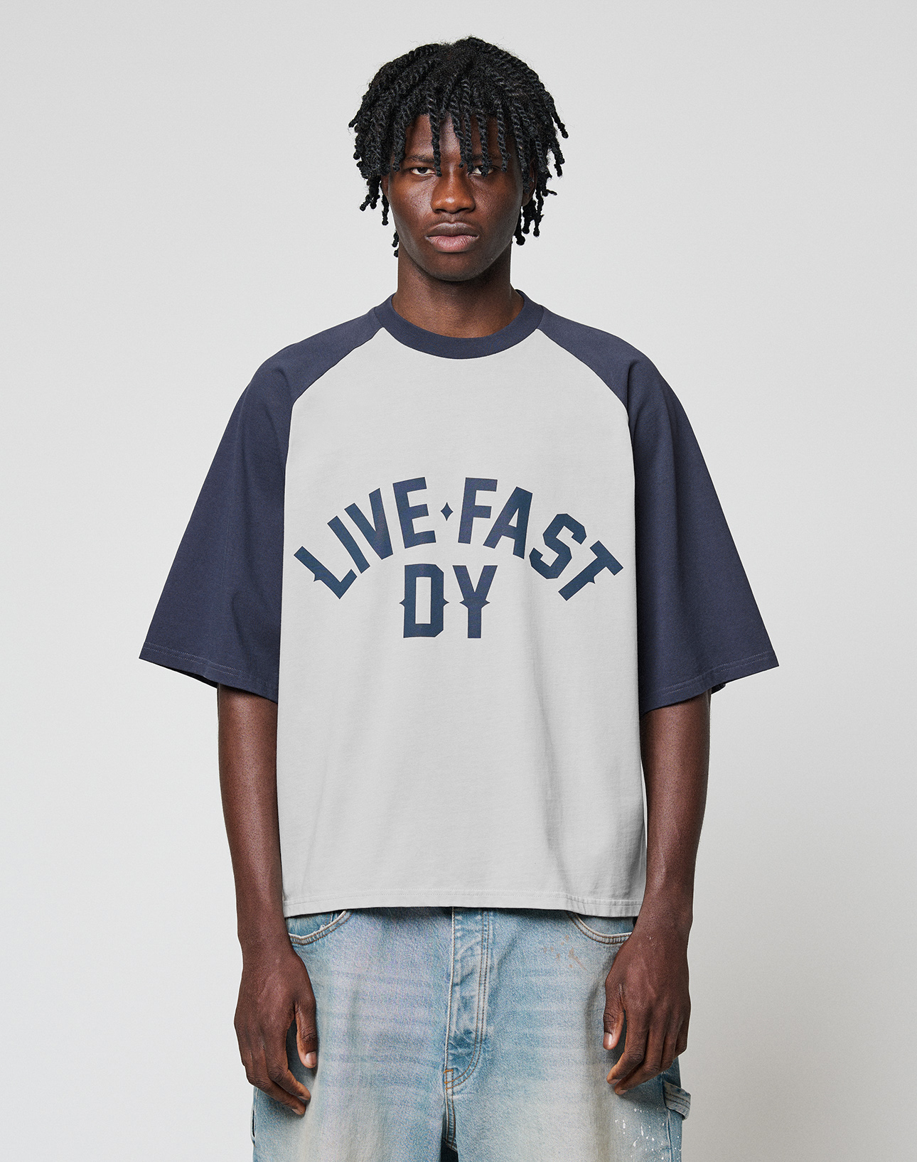 A person faces the camera in a loose-fit, two-tone Live Fast Raglan Tee by LIVE FAST DIE YOUNG with bold lettering on the front, paired with light blue jeans against a plain light backdrop. | model