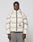 A person in an oversize, light beige Live Fast Puffer Jacket by LIVE FAST DIE YOUNG with a hood, front zipper, and reflective "LIVE FAST" logo stands facing forward. They have short braided hair and wear wide gray pants.