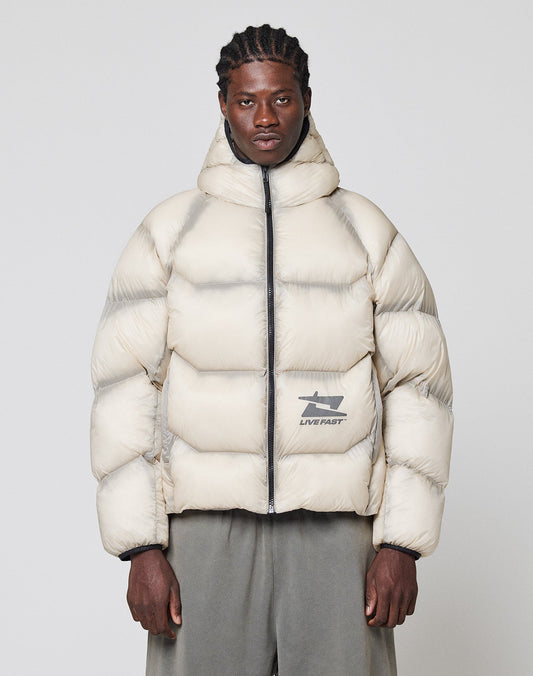 A person in an oversize, light beige Live Fast Puffer Jacket by LIVE FAST DIE YOUNG with a hood, front zipper, and reflective "LIVE FAST" logo stands facing forward. They have short braided hair and wear wide gray pants.