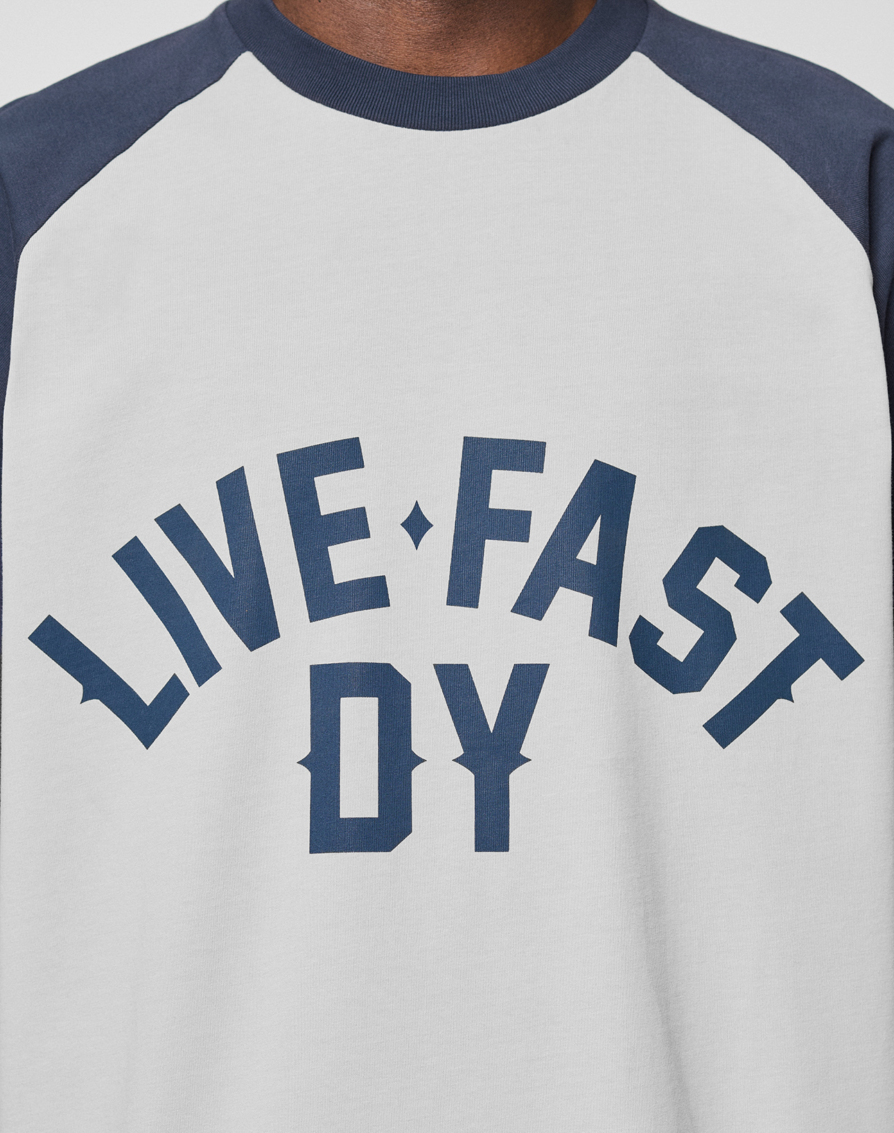 Close-up of a person wearing the Live Fast Raglan Tee by LIVE FAST DIE YOUNG, a boxy oversize fit white shirt with dark raglan sleeves and bold navy "LIVE FAST DY" text, with "DY" centered below "LIVE FAST.