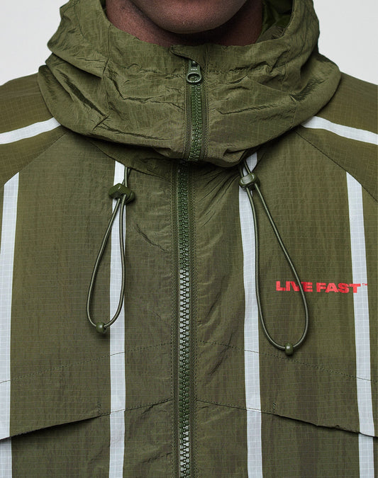 Close-up of a person wearing the olive green LFDY Nylon Trackjacket by LIVE FAST DIE YOUNG, featuring an oversize fit, Kapuze mit Elastik-Tunnelzug, white vertical stripes, red LIVE FAST chest text, zip-up front, and Front- und Backprint.