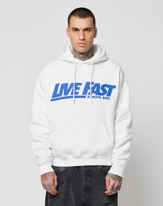 A person with short hair and visible tattoos wears the LIVE FAST DIE YOUNG Live Fast Europe Run Hooded, a white oversize hoodie with blue text, Kapuze, standing facing forward against a plain light background.