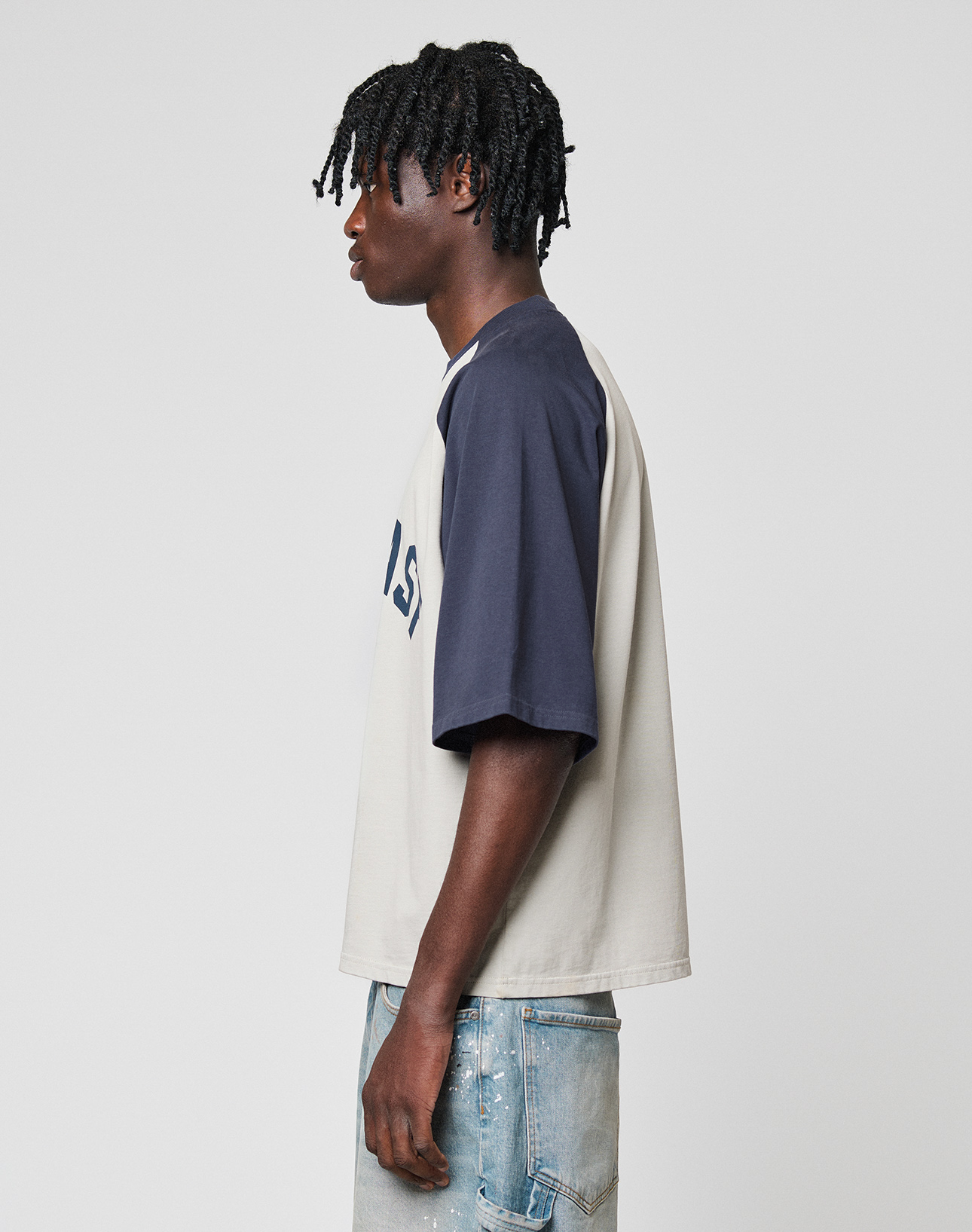 A person with medium brown skin and short dreadlocks stands in profile, wearing the LIVE FAST DIE YOUNG Live Fast Raglan Tee in beige and navy with oversized fit, paired with faded blue jeans against a plain light background.