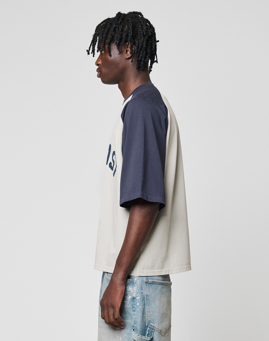 A person with medium brown skin and short dreadlocks stands in profile, wearing the LIVE FAST DIE YOUNG Live Fast Raglan Tee in beige and navy with oversized fit, paired with faded blue jeans against a plain light background.