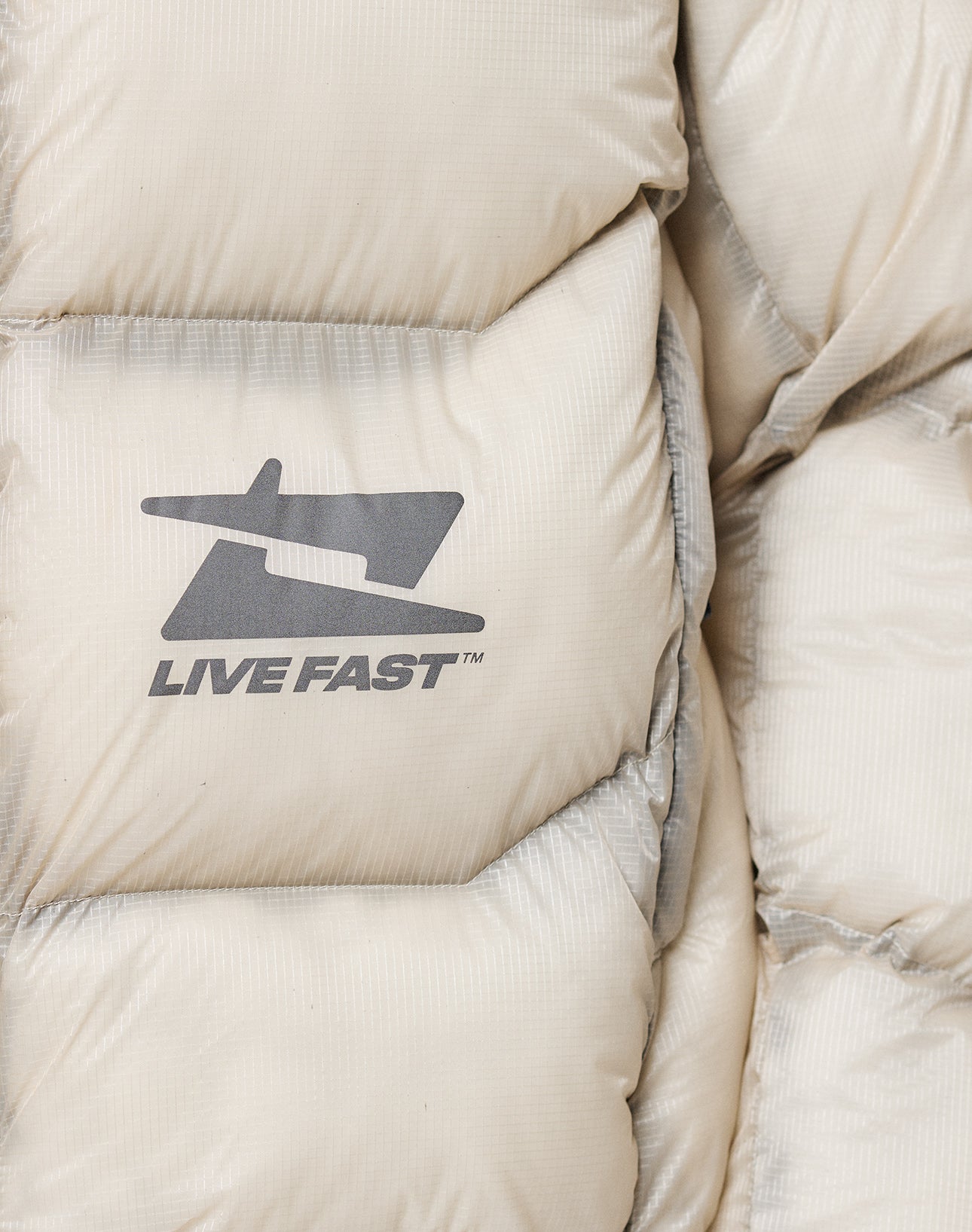 Close-up of the Live Fast Puffer Jacket by LIVE FAST DIE YOUNG, an oversize light beige puffer with quilted stitching, a stylized airplane logo, and LIVE FAST™ in dark gray reflektierender Print on durable nylon fabric.
