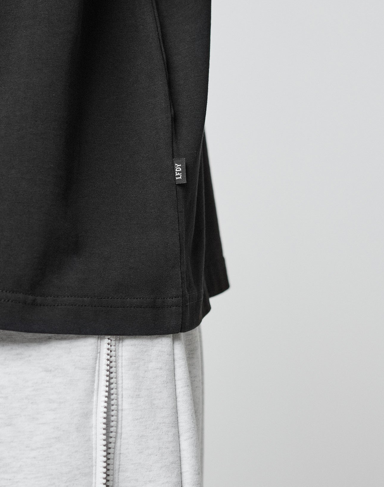 Close-up of the LFDY Paris Tee by LIVE FAST DIE YOUNG in an oversize black fit with a small side tag, paired with light gray pants featuring a visible vertical zipper. The background is plain and light-colored.