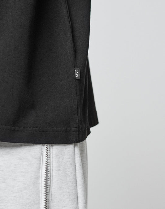 Close-up of the LFDY Paris Tee by LIVE FAST DIE YOUNG in an oversize black fit with a small side tag, paired with light gray pants featuring a visible vertical zipper. The background is plain and light-colored.