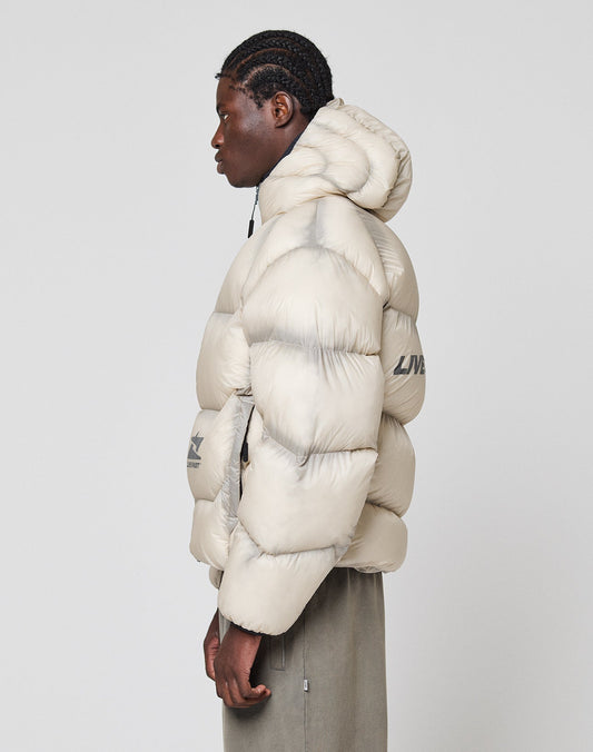 A person with braided hair, in profile, wears the LIVE FAST DIE YOUNG Live Fast Puffer Jacket in oversize light beige nylon with a hood. Reflective print and subtle logo details accent the sleeve and chest.