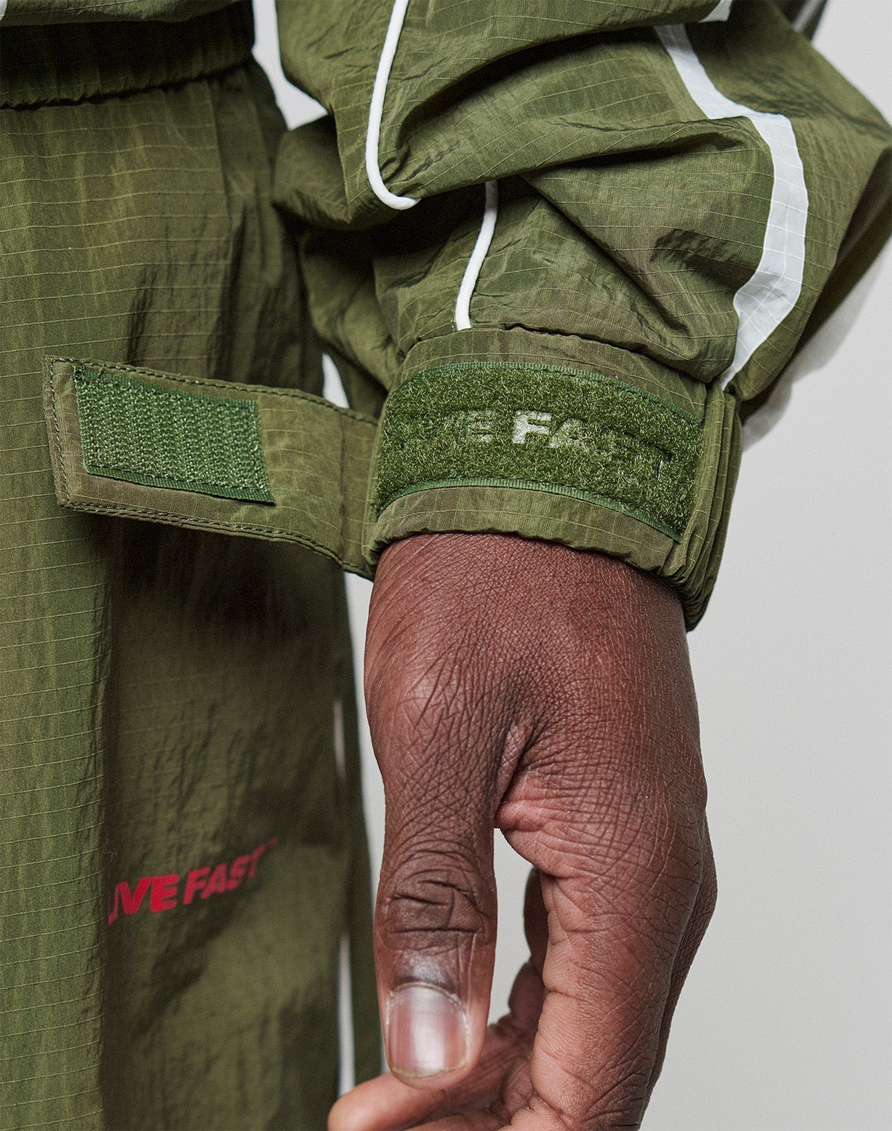 A close-up of a hand adjusting the green Velcro cuff on the olive LFDY Nylon Trackjacket by LIVE FAST DIE YOUNG, featuring white stripes, a textured pattern, partial “IVE FAST” text on the sleeve, and a hood with elastic drawcord.
