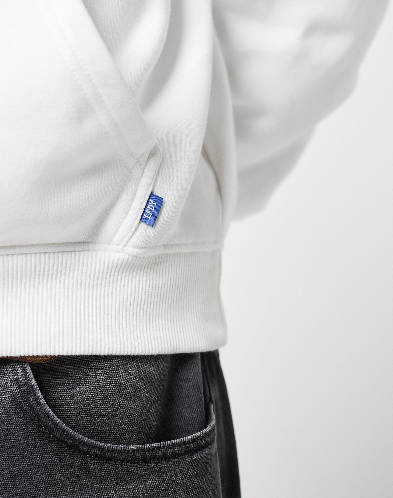 Close-up of a person wearing the Live Fast Europe Run Hooded sweatshirt by LIVE FAST DIE YOUNG in white Baumwolle with an oversize fit, showing a small blue tag and black jeans, focusing on the lower side of the sweatshirt and upper jeans.