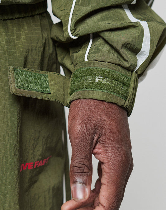 A close-up of a hand adjusting the green Velcro cuff on the olive LFDY Nylon Trackjacket by LIVE FAST DIE YOUNG, featuring white stripes, a textured pattern, partial “IVE FAST” text on the sleeve, and a hood with elastic drawcord.