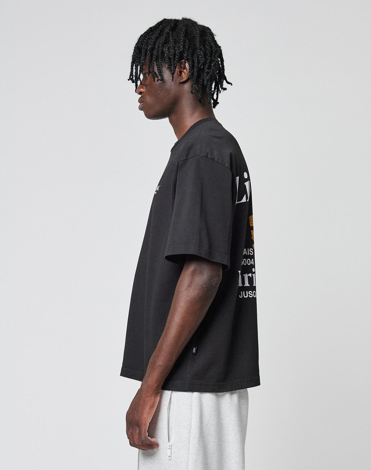 A young man with short dreadlocks stands in profile against a plain light gray background, wearing the LIVE FAST DIE YOUNG LFDY Paris Tee—an oversize fit black graphic t-shirt with front and back print—and light gray shorts.