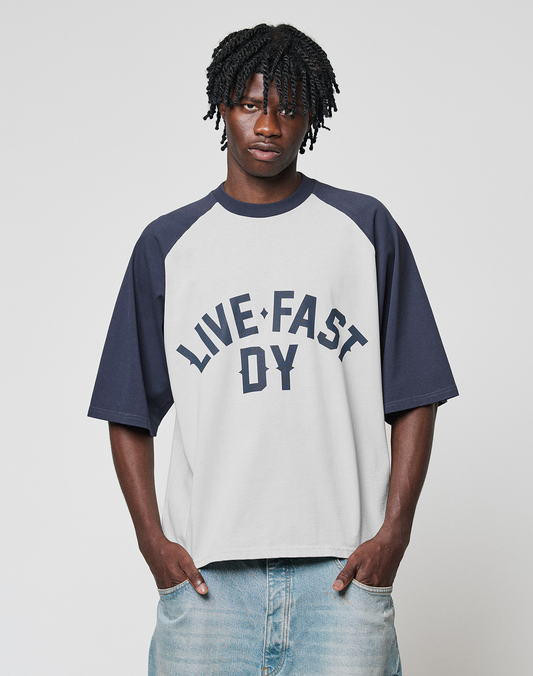 A man with braided hair wears the Live Fast Raglan Tee by LIVE FAST DIE YOUNG—an oversized white and dark blue raglan t-shirt. He stands with hands in his pockets, looking at the camera with a neutral expression.