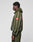 A person stands sideways wearing the LIVE FAST DIE YOUNG LFDY Nylon Trackjacket in green with a relaxed fit, white vertical stripes, hood with elastic drawstring, and bold red patches with white logos on the sleeve and back. Plain light gray background.