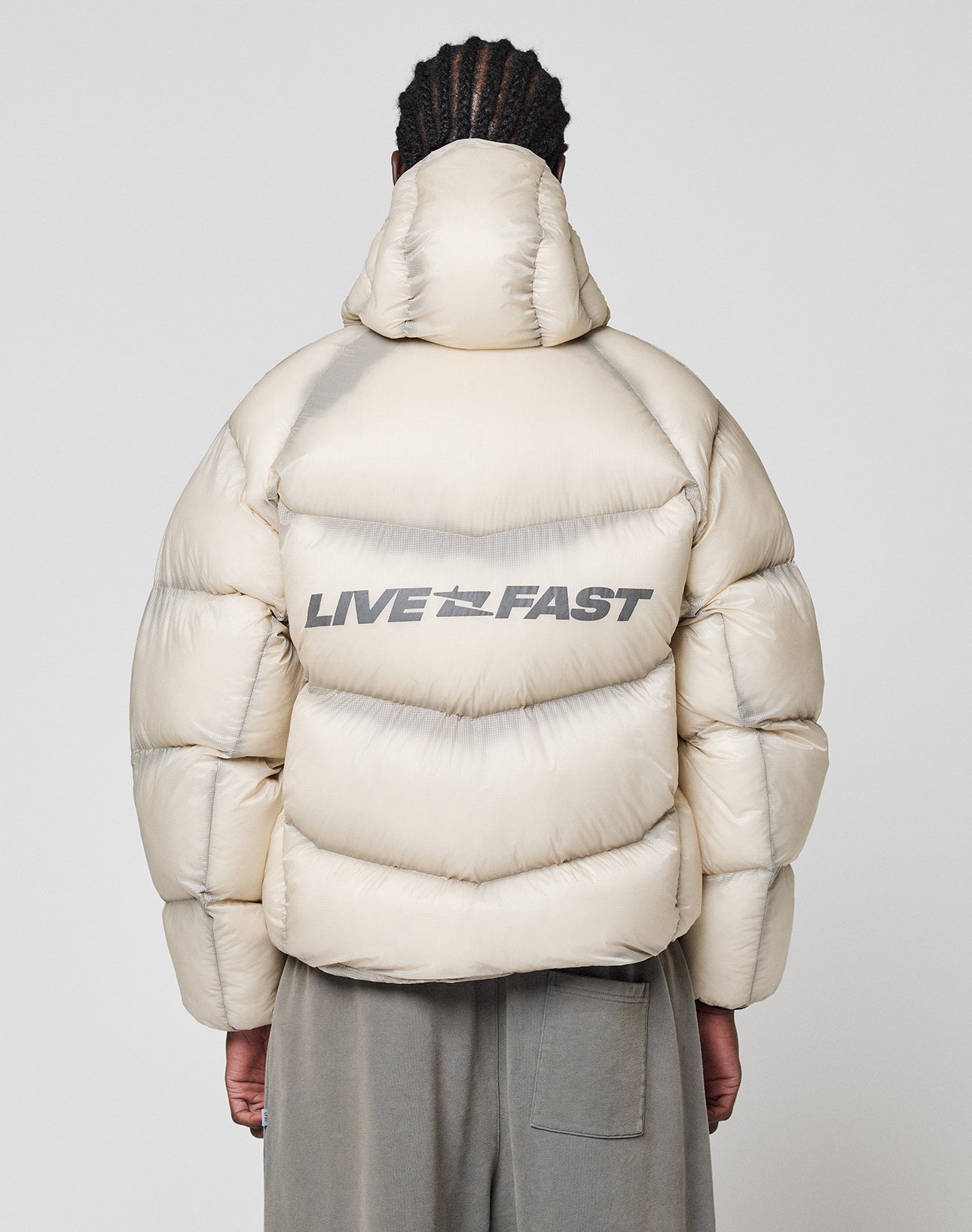 A person faces away from the camera, wearing a light beige LIVE FAST DIE YOUNG "Live Fast Puffer Jacket" with an oversize fit and bold reflective "LIVE FAST" print on the back, standing against a plain white background.