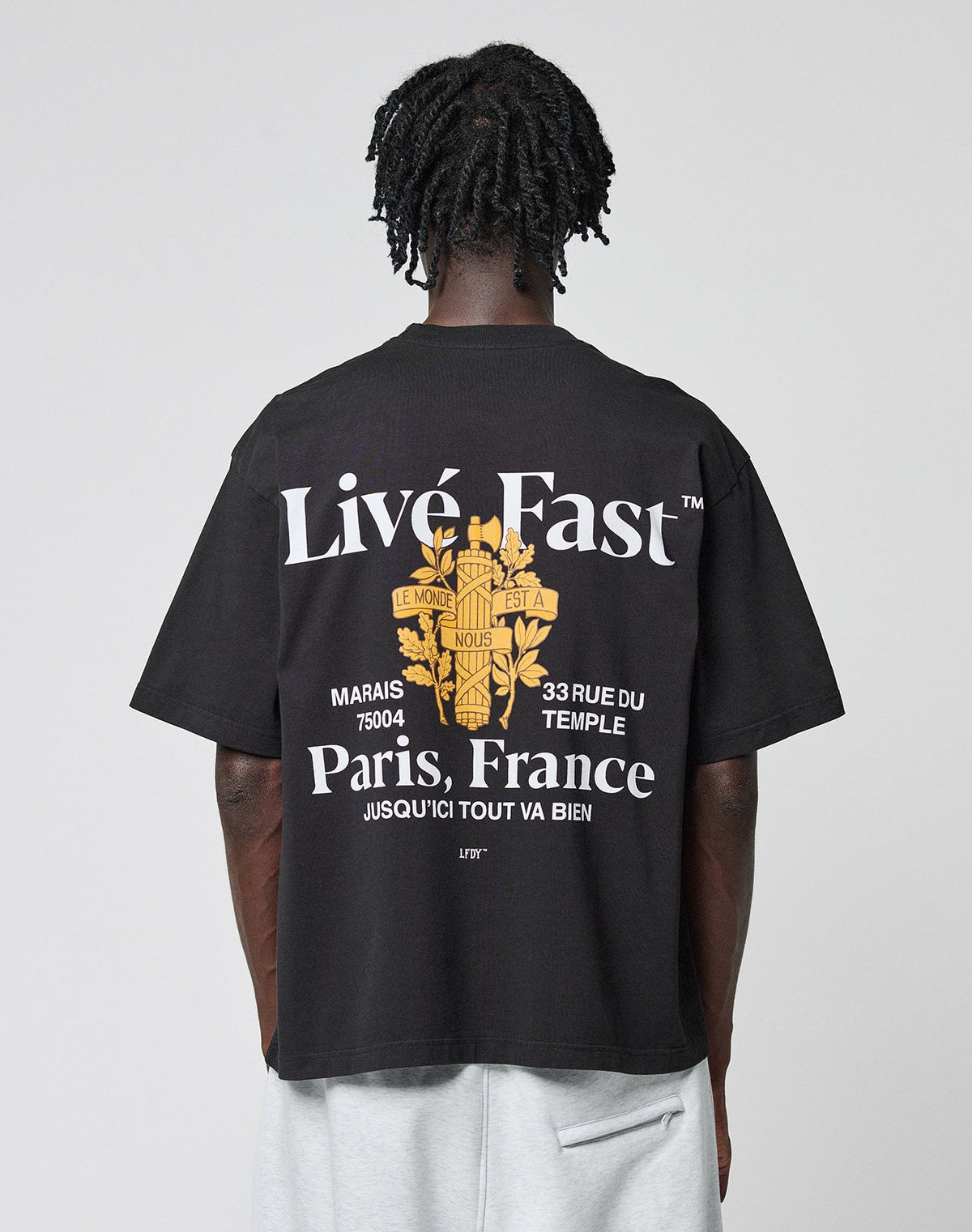 A person with short, twisted hair faces away, wearing the LIVE FAST DIE YOUNG LFDY Paris Tee in Oversize Fit. The black shirt features bold white text, a yellow Paris graphic, French phrases, a floral design, and prominent front and back prints. | model