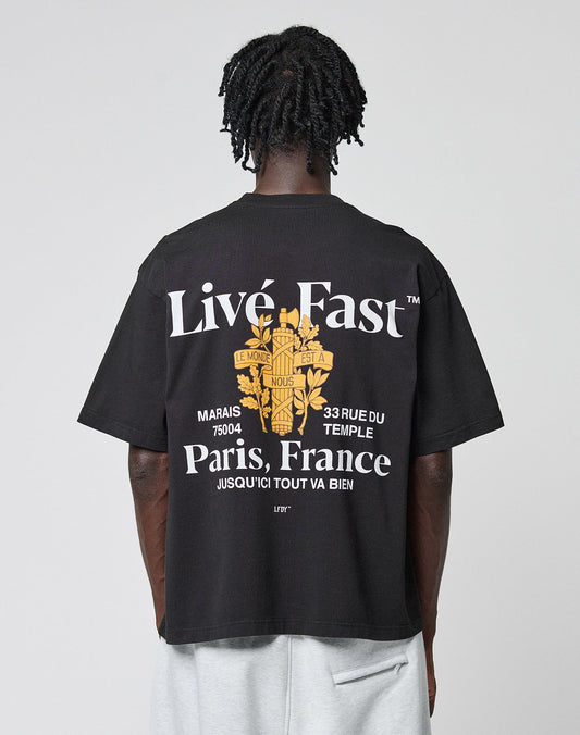 A person with short, twisted hair faces away, wearing the LIVE FAST DIE YOUNG LFDY Paris Tee in Oversize Fit. The black shirt features bold white text, a yellow Paris graphic, French phrases, a floral design, and prominent front and back prints. | model