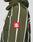 A person wears the LFDY Nylon Trackjacket by LIVE FAST DIE YOUNG—an olive green, oversize jacket with white vertical stripes, a red patch with a white geometric symbol on the sleeve, visible stitching, and a hood with elastic tunnelzug.