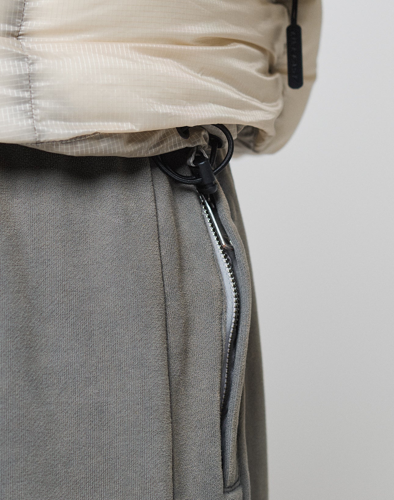 Close-up of a person in grey sweatpants and the Live Fast Puffer Jacket by LIVE FAST DIE YOUNG, featuring an oversize fit in light beige and a hem with a black adjustable drawstring and toggle.